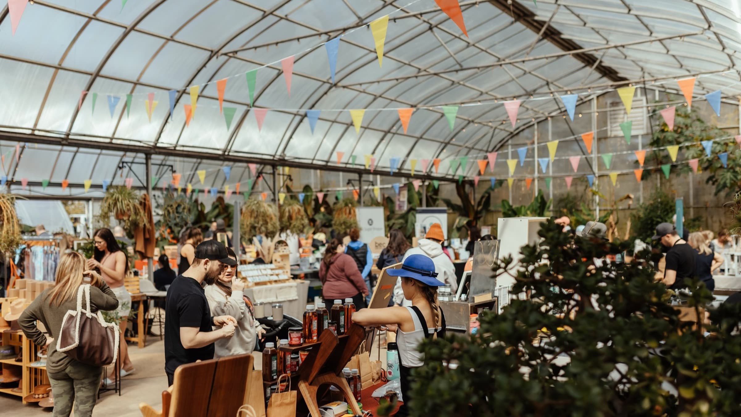 A market running under a greenhouse roof, bunting strung across it, shoppers moving between the stalls